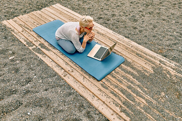 Positive well-fit blond woman in sportswear, seated in lotus pose, using laptop at the beach....