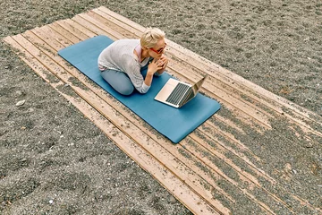 Tuinposter Positive well-fit blond woman in sportswear, seated in lotus pose, using laptop at the beach. Freelance working, wellbeing concept. © Caterina Trimarchi
