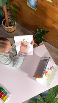 Creative broadcasting, childhood hobby. Vertical of little kid blogger shares drawing tips on digital tablet indoor. Inspiring online art channel for children on backdrop of pictures and houseplants
