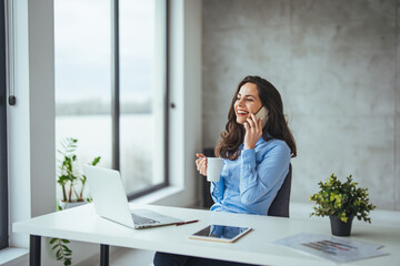 Young businesswoman sitting at her desk talking on phone and working on computer. Female executive working in modern workplace. Young businesswoman using mobile phone and laptop