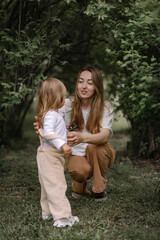 A small stylish girl, a child and her mother sat down on a green lawn in a park among bushes. Mom hugs her daughter. The daughter is holding a basket and a branch of a blossoming apple tree.