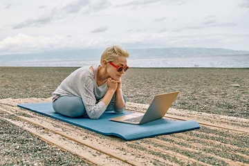 Tuinposter Positive well-fit blond woman in sportswear, seated in lotus pose, using laptop at the beach. Freelance working, wellbeing concept. © Caterina Trimarchi