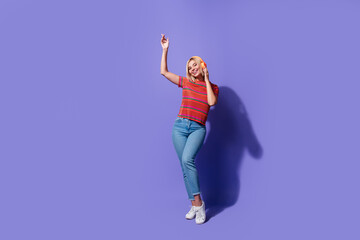 Full body photo of carefree blonde lady in striped t shirt denim jeans likes modern bluetooth earphones isolated on violet color background