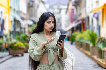 Shocked and disappointed young Indian student girl standing on city street and looking at mobile phone screen pointing with hand - obrazy, fototapety, plakaty