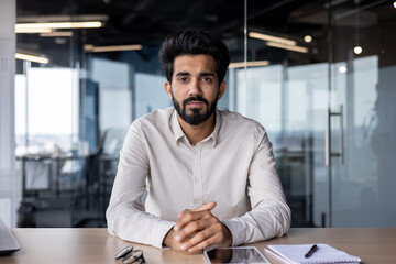 Portrait of a successful and serious Indian male businessman sitting at the office desk and confidently looking at the camera