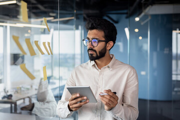 Serious and focused young Indian man working in office with tablet, standing near glass board with...