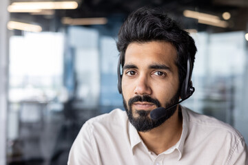 Serious Indian man wearing a headset works in the office and looks confidently at the camera,...