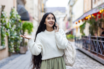 Fototapeta premium An Indian young smiling woman walks around the city, talks expressively on a mobile phone, has an emotional discussion, gestures with her hands