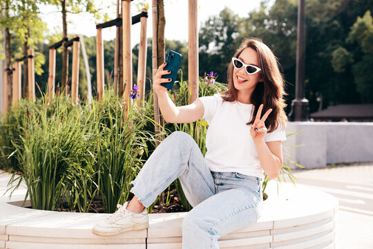 Young beautiful smiling hipster woman in trendy summer white t-shirt and jeans clothes. Sexy carefree model posing on the street background at sunset. Positive model takes selfie photos. In sunglasses