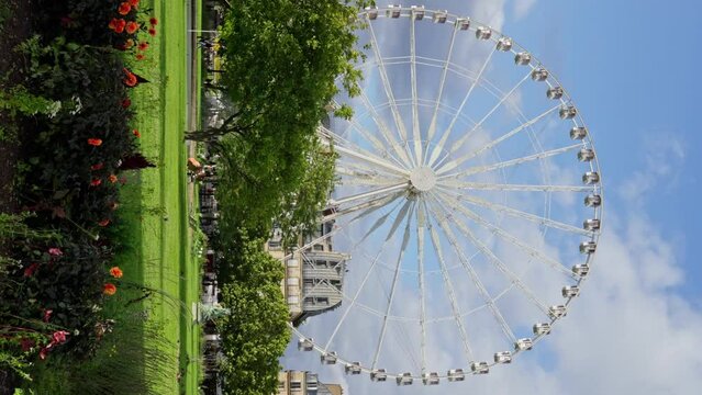 A sky-high ferris wheel surrounded by water, clouds, plants, and electric blue sky in a park of recreation. Paris. France