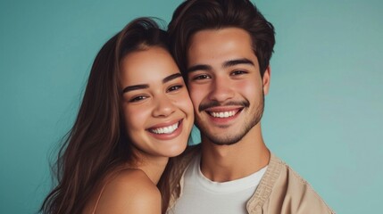 A young couple sharing a joyful moment smiling brightly at the camera with their heads close together against a soft teal background showcasing a sense of closeness and happiness.