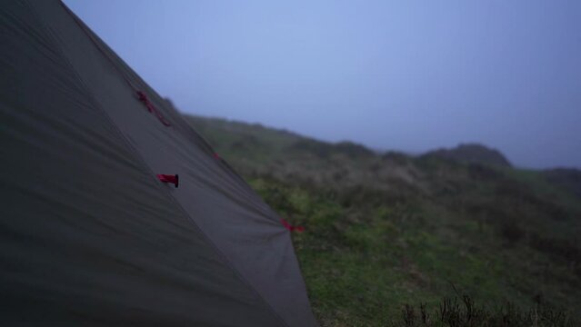 A grim dull camping scene with a tent flapping in bad weather on a hillside