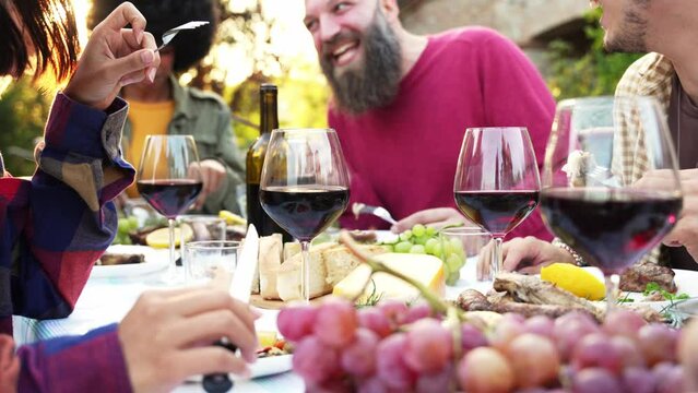 Happy friends having bbq dinner party in garden restaurant - Cheerful family drinking red wine together sitting at dining table - Food and beverage concept with guys and girls eating fresh meal
