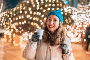 A woman enjoys the winter streets, sipping mulled wine amidst the festive glow of Christmas garlands.