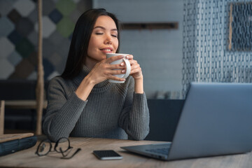 Young female manager drinking with pleasure coffee during break sitting and relax at workplace in...