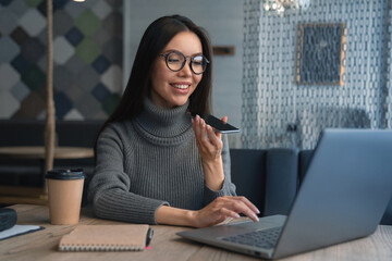 Happy business woman talking on speakerphone sitting at table with laptop, paper cup for coffee and notebook on it. Asian female sitting at office having telephonic conversation and smiling
