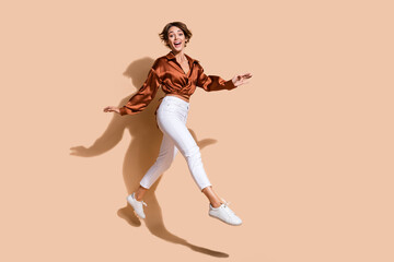 Fototapeta na wymiar Full length photo of excited pretty woman wear brown blouse jumping high walking empty space isolated beige color background