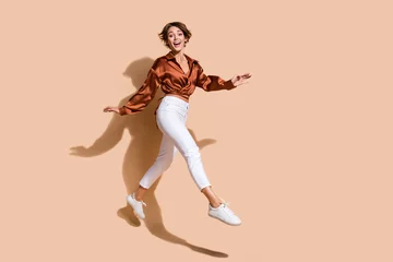 Tapeten Full length photo of excited pretty woman wear brown blouse jumping high walking empty space isolated beige color background © deagreez