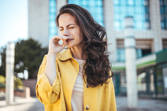 Woman with allergy symptom blowing nose. Outdoor shot of displeased Caucasian woman feels allergy, holds white tissuue, stands near tree with blossom, feels unwell, sneezes all time. 