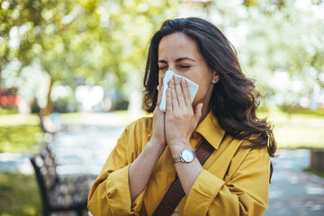 Shot of an attractive young woman feeling ill and blowing her nose with a tissue outdoors. Woman has sneezing. Young woman is having flu and she is sneezing. Sickness, seasonal virus problem concept - obrazy, fototapety, plakaty