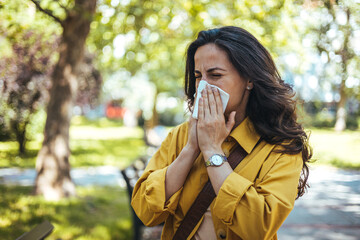 Portrait of unhealthy cute  female in yellow top with napkin blowing nose, looks to the source of the allergy, place for advertising. Rhinitis, cold, allergy concept. Pollen allergy symptoms - obrazy, fototapety, plakaty