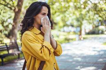 Portrait of unhealthy cute  female in yellow top with napkin blowing nose, looks to the source of the allergy, place for advertising. Rhinitis, cold, allergy concept. Pollen allergy symptoms - obrazy, fototapety, plakaty