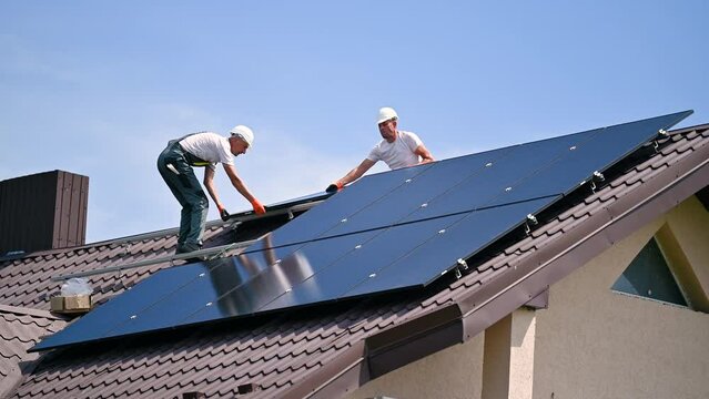 Workers building solar panel system on roof of house. Two men installers in helmets carrying photovoltaic solar module outdoors. Alternative, green and renewable energy generation concept.