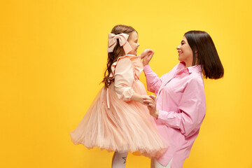 Fototapeta premium Birthday celebration. Stylish happy mother dancing with her little pretty daughter, kid against yellow studio background. Concept of happiness, Mother's day, childhood, fashion and lifestyle