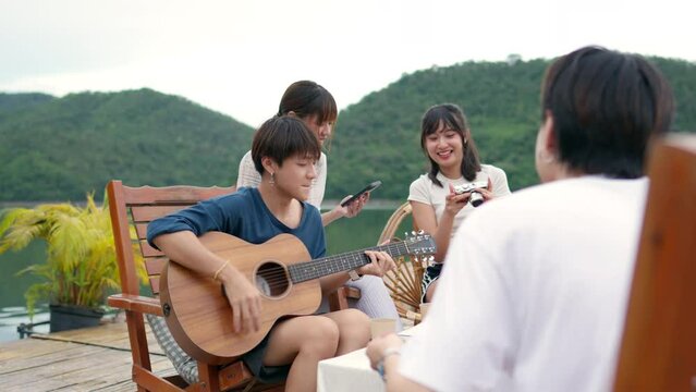 Young Asian man and woman enjoy outdoor lifestyle travel nature forest mountain on summer holiday vacation. Generation z people friends party playing guitar and singing together at lake house balcony.