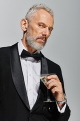 good looking bearded mature man in elegant tuxedo holding glass of martini and looking at camera