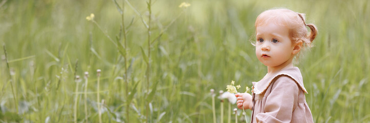 A child in a light brown dress stands in a field, holding a small flower. Surrounded by greenery, the scene captures a moment of innocence and serenity. Banner with copy space.