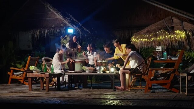 Group of Diversity Asian man and woman friends enjoy and fun celebration meeting dinner party eating barbecue grill and drinking beer together on lake house balcony on summer holiday vacation at night