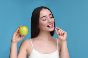 Beautiful young woman with vitamin pill and apple on light blue background