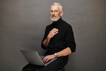 appealing mature man in trendy black turtleneck sitting on chair with laptop and looking at camera - 731004838