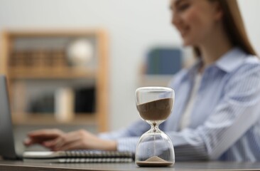 Hourglass with flowing sand on desk. Woman using laptop indoors, selective focus