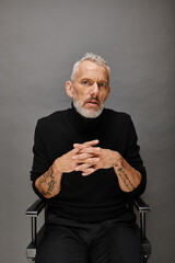 attractive mature man with beard in trendy turtleneck sitting on folding chair and looking at camera