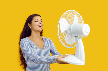 Summer heat. Joyful woman enjoys flow of air from an electric fan isolated on yellow background....