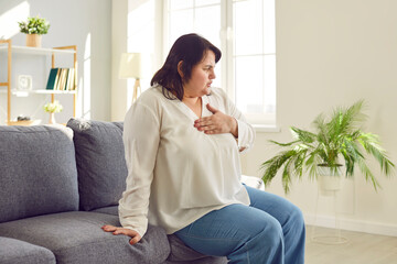 Overweight young woman clutching her chest suffering from pain. Plus size woman sitting on sofa st...