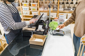 Woman paying with contactless credit card with NFC technology in local zero waste grocery store at cash desk. Shelf filled with bulk organic food on background. Green shopping concept