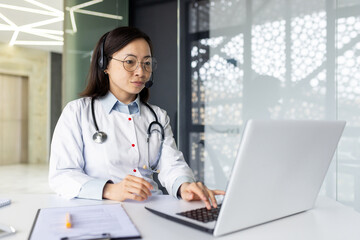 Experienced Asian doctor offers telemedicine services, remotely consulting patients from a clinic office with a headset and laptop.