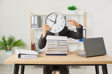 Overworked funny business man hiding head behind the clock at his workplace with a pile of folders...