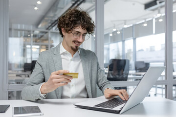 Confident young male entrepreneur with curly hair engaged in online financial operations, using a credit card and laptop at his workplace.