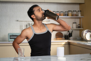 Happy Middle aged fit man drinking protein shake from bottle at kitchen after morning workout -...