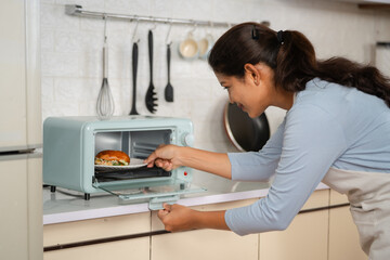 Indian happy smiling woman cooking burger by placing it on microwave oven at kitchen - concept of...
