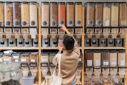 African American woman refilling reusable package with bulk products in local zero waste grocery store. Rear view of woman buying grains and cereals in sustainable eco grocery store.