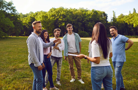 Group of happy friends or corporate coworkers standing on grass in summer park on weekend and listening to manager with clipboard listing their tasks and explaining rules of outdoor team building game