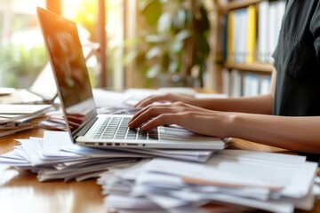At the desk working person on laptop computer with a pile of documents on table in office. Overwork and excess responsibilities at work.