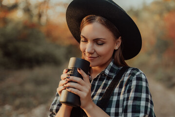 Hipster woman enjoys aroma and taste of fresh coffee. Drinking from thermos in autumn forest