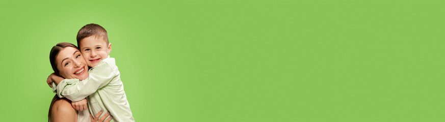 Happy, smiling little boy, son hugging his beautiful, caring mother, showing his love against green studio background. Concept of happiness, Mother's day, childhood, fashion and lifestyle