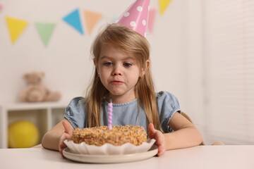 Cute girl in party hat with birthday cake at table indoors